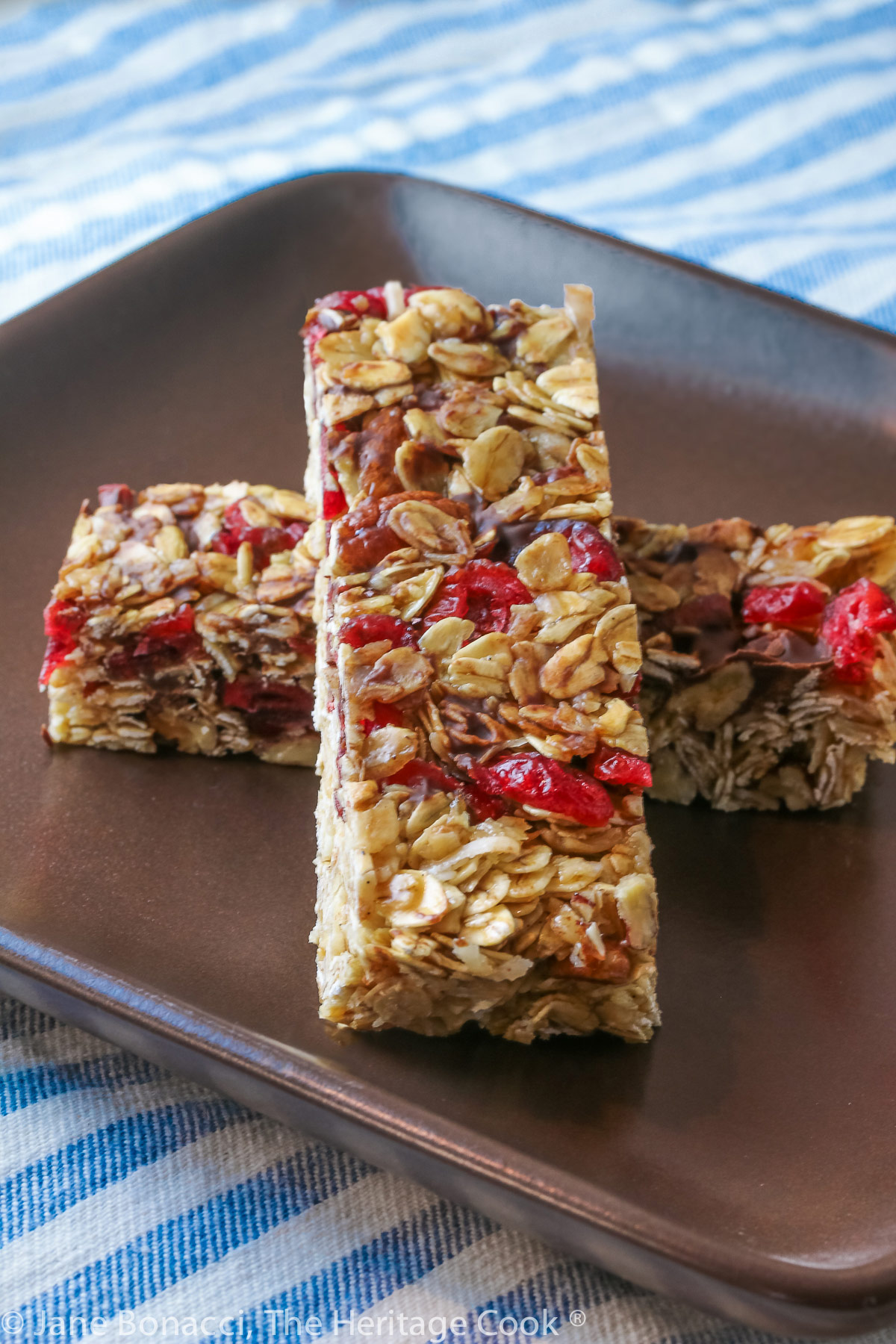 Two granola bars studded with red cranberries and chocolate set perpendicular to each other one on top in the shape of an X on a dark brown square plate © 2023 Jane Bonacci, The Heritage Cook. 