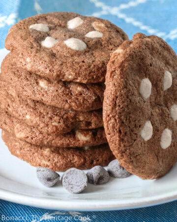 A stack of 5 dark chocolate cookies with white chocolate chips arranged on the top of each cookie, some with a cookie propped up against the stack, all on a white plate; Quadruple Chocolate Cookies © 2023 Jane Bonacci, The Heritage Cook.