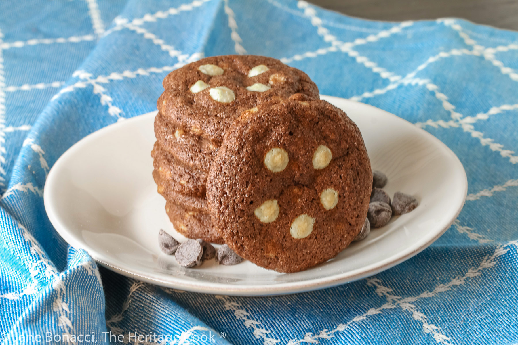 A stack of 5 dark chocolate cookies with white chocolate chips arranged on the top of each cookie, some with a cookie propped up against the stack, all on a white plate; Quadruple Chocolate Cookies © 2023 Jane Bonacci, The Heritage Cook. 