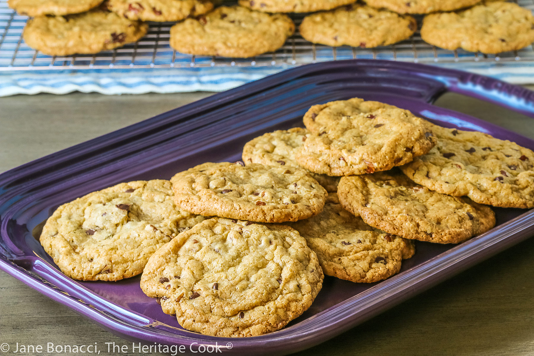 Golden brown cookies studded with chocolate, caramel, and pecans piled on a wire rack and purple platter; Turtle Chocolate Chip Cookies © 2023 Jane Bonacci, The Heritage Cook. 