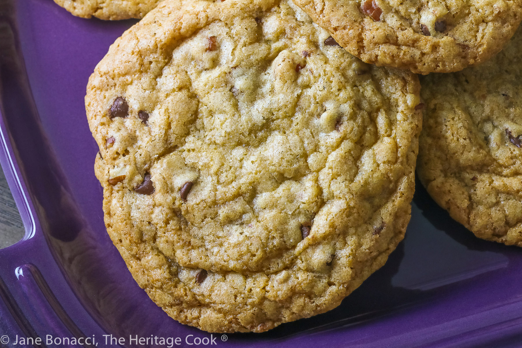 Close up of a single cookie; Golden brown cookies studded with chocolate, caramel, and pecans piled on a wire rack and purple platter; Turtle Chocolate Chip Cookies © 2023 Jane Bonacci, The Heritage Cook. 