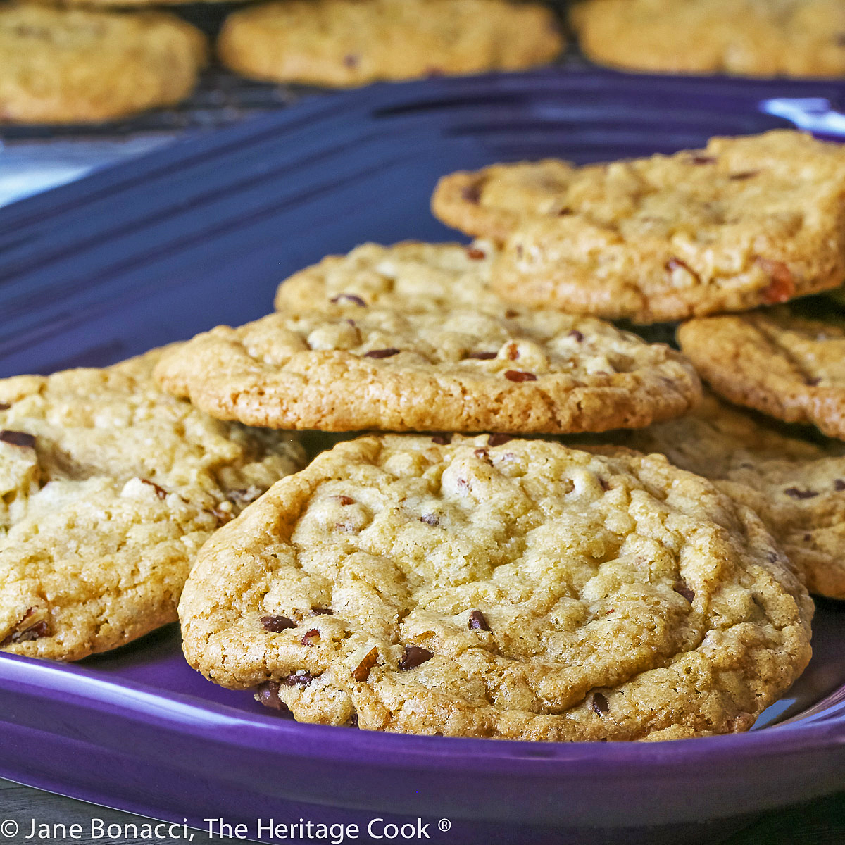 Golden brown cookies studded with chocolate, caramel, and pecans piled on a wire rack and purple platter; Turtle Chocolate Chip Cookies © 2023 Jane Bonacci, The Heritage Cook. 