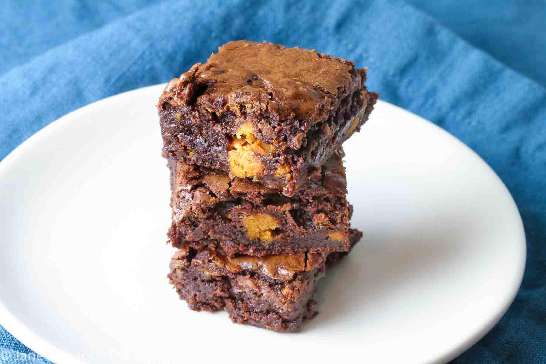 Butterfinger Brownies stacked on white plate with teal napkin background, one with a hand reaching for a brownie off the stack © 2023 Jane Bonacci, The Heritage Cook.