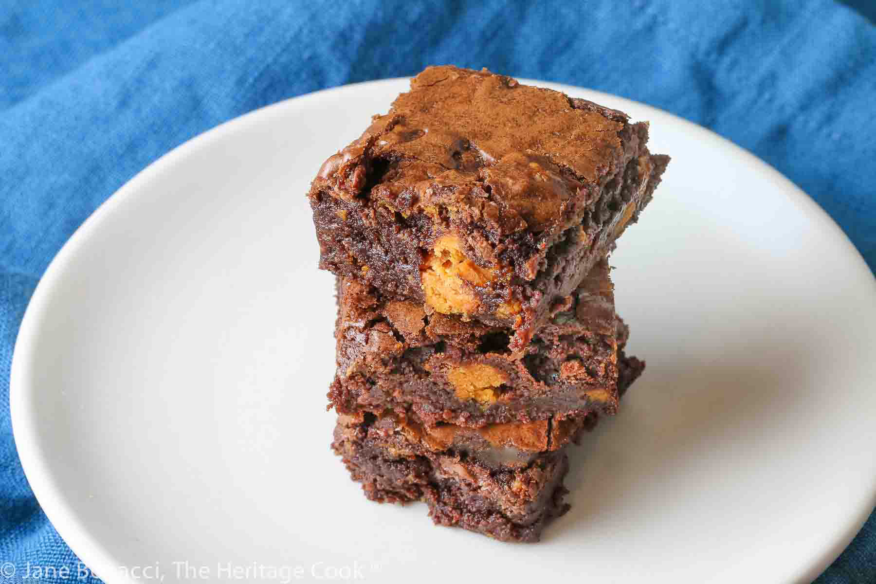 Butterfinger Brownies stacked on white plate with teal napkin background, one with a hand reaching for a brownie off the stack © 2023 Jane Bonacci, The Heritage Cook.