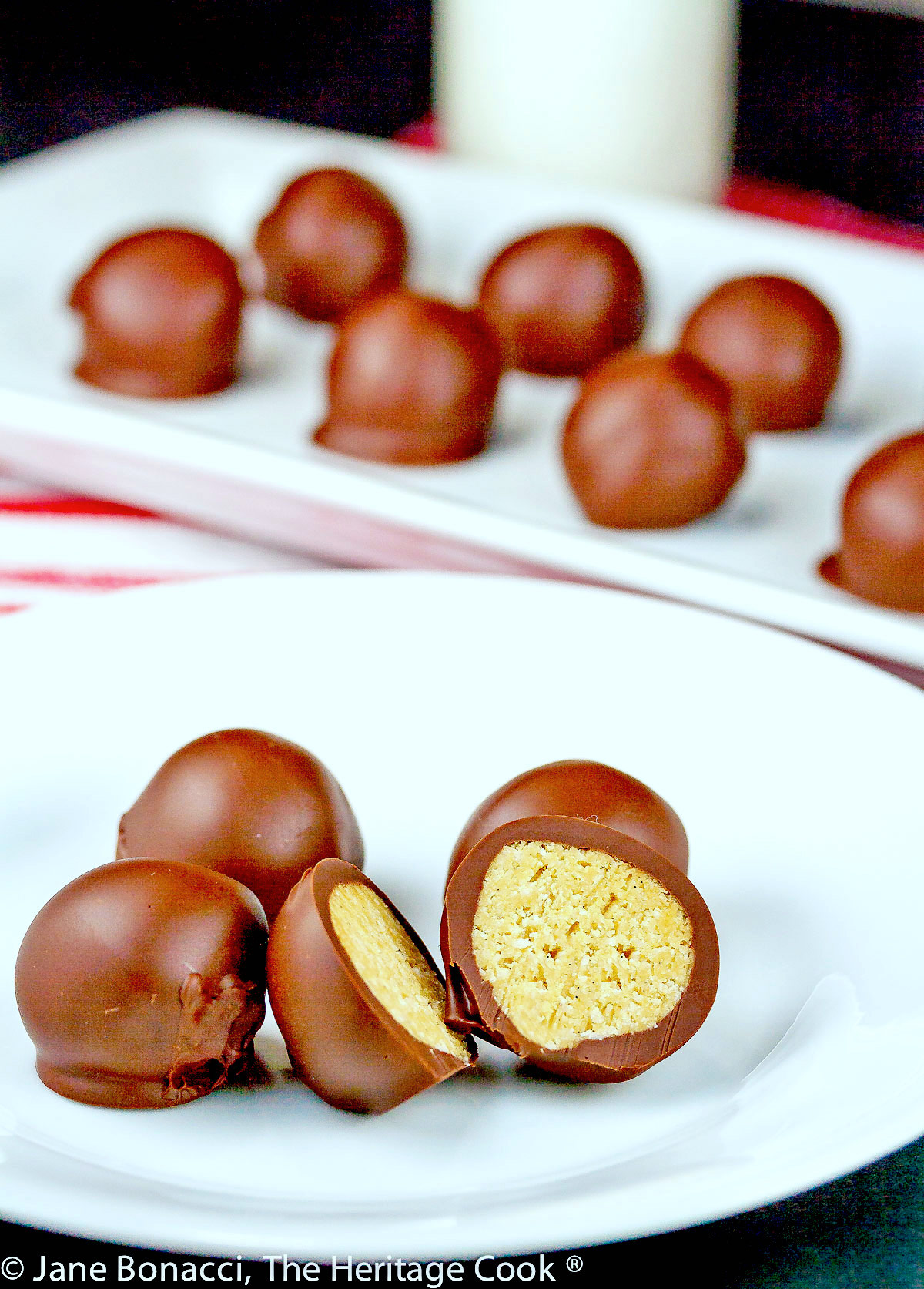 Chocolate Peanut Butter Truffles are balls of sweetened peanut butter filling coated in dark chocolate, some cut open to show the centers, on a white tray or plate on top of red striped towels © 2023 Jane Bonacci, The Heritage Cook. 