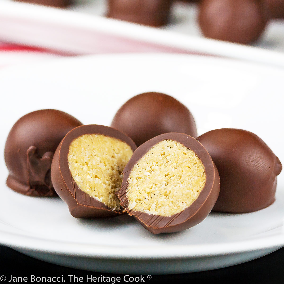 Chocolate Peanut Butter Truffles are balls of sweetened peanut butter filling coated in dark chocolate, some cut open to show the centers, on a white tray or plate on top of red striped towels © 2023 Jane Bonacci, The Heritage Cook.