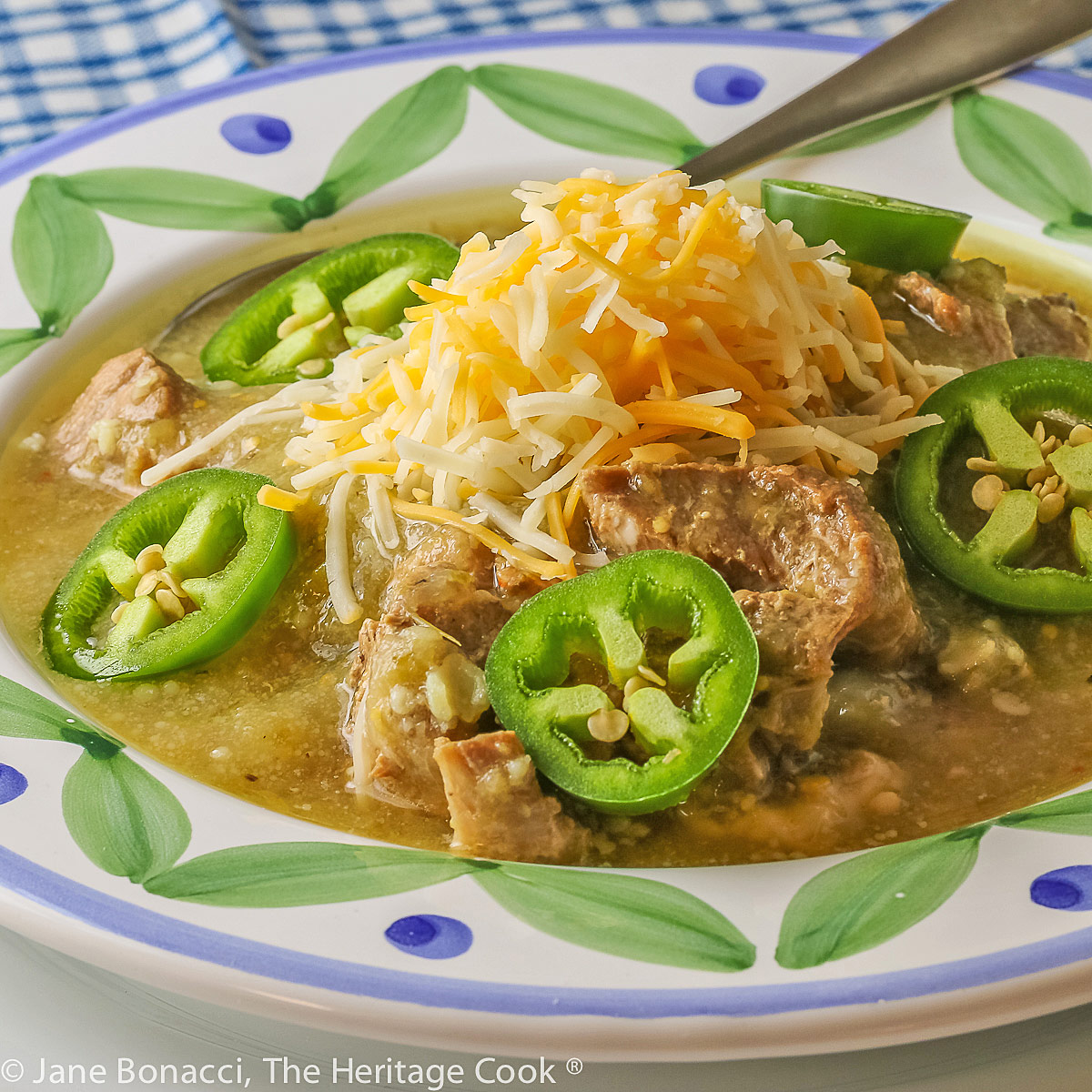 Colorful decorated bowl filled to the rim with Pork Chile Verde, topped with cheese, sliced jalapenos, and cilantro © 2023 Jane Bonacci, The Heritage Cook.