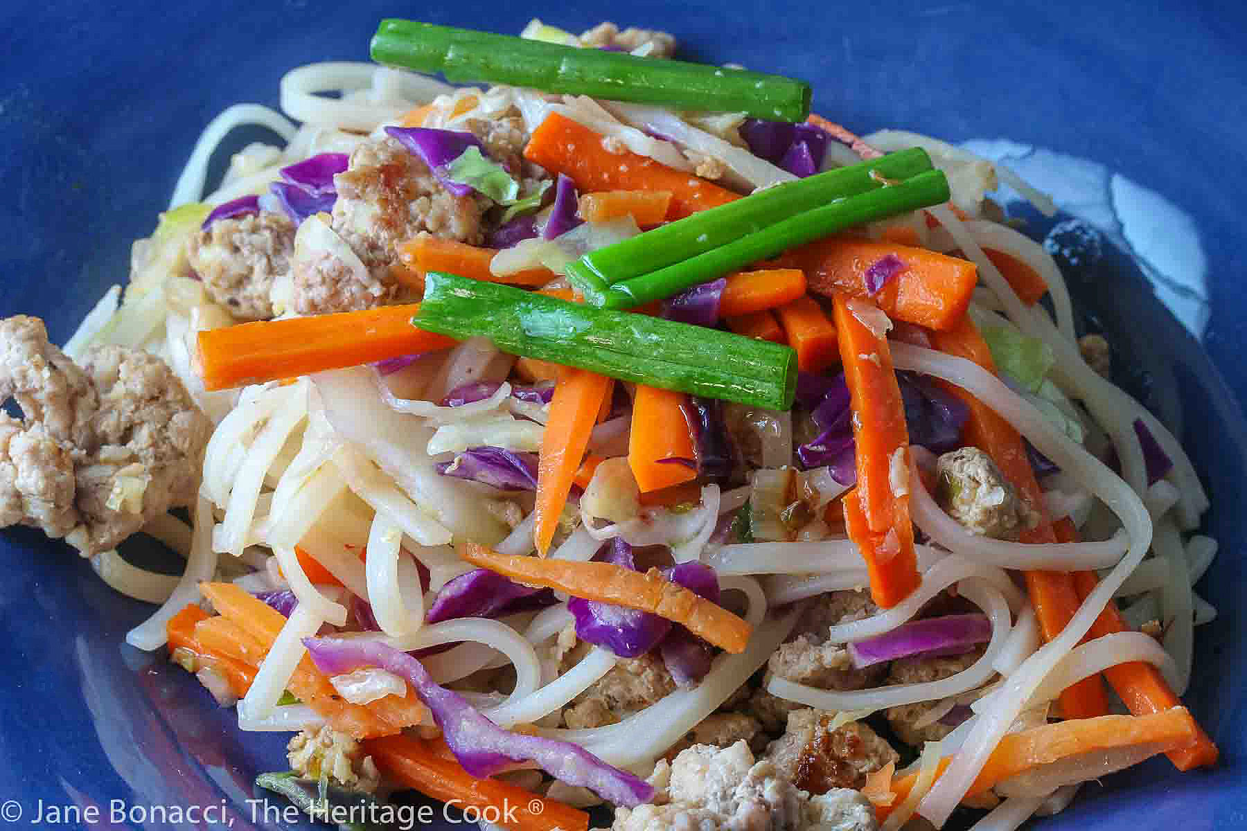 Big serving of noodles, pork, and bright vegetables in a bold blue bowl with chopsticks in the back, sitting on a colorful plaid background © 2023 Jane Bonacci, The Heritage Cook. 