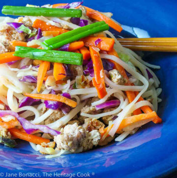 Big serving of noodles, pork, and bright vegetables in a bold blue bowl with chopsticks in the back, sitting on a colorful plaid background © 2023 Jane Bonacci, The Heritage Cook.
