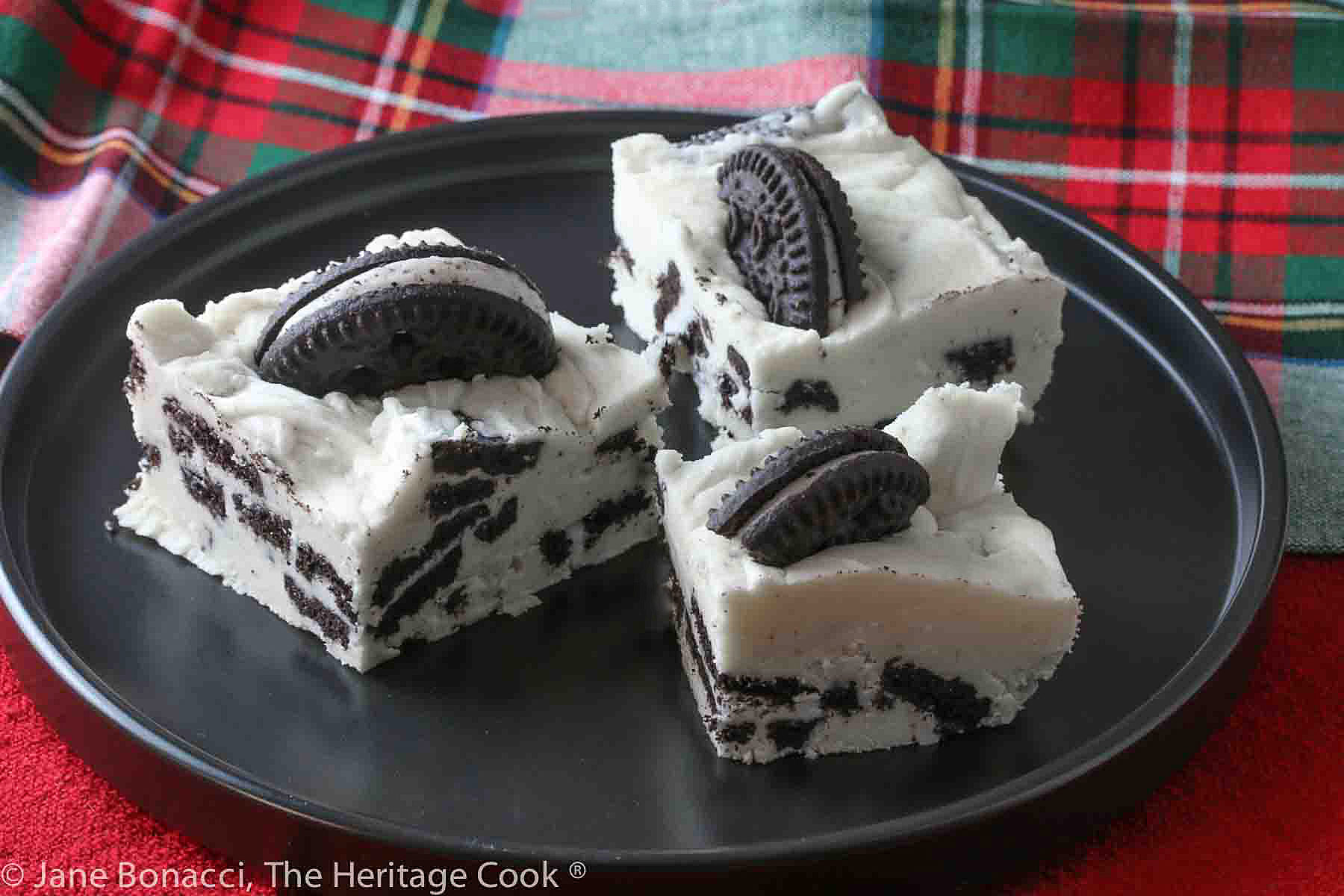 3 Squares of Cookies and Cream Fudge on a black plate on top of red and holiday plaid cloths © 2023 Jane Bonacci, The Heritage Cook. 