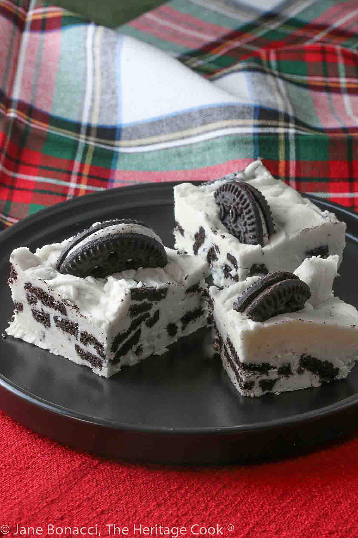 3 Squares of Cookies and Cream Fudge on a black plate on top of red and holiday plaid cloths © 2023 Jane Bonacci, The Heritage Cook. 