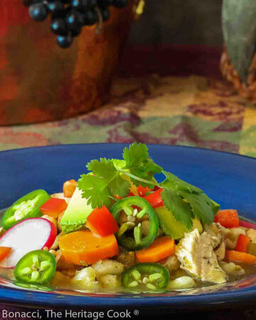 Large bowl of green posole turkey soup topped with your choice of garnishes, sitting on a festive harvest colored cloth; I used cilantro, limes, avocado, red bell pepper, and radishes for brightness and interest © 2023 Jane Bonacci, The Heritage Cook.