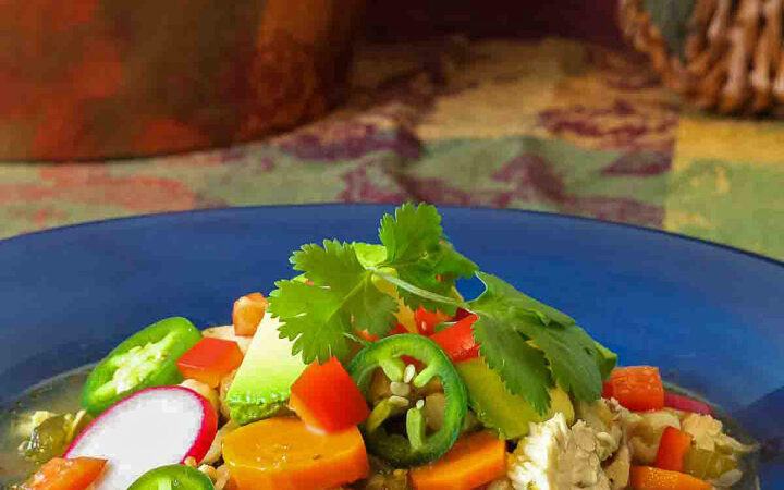 Large bowl of green posole turkey soup topped with your choice of garnishes, sitting on a festive harvest colored cloth; I used cilantro, limes, avocado, red bell pepper, and radishes for brightness and interest © 2023 Jane Bonacci, The Heritage Cook.