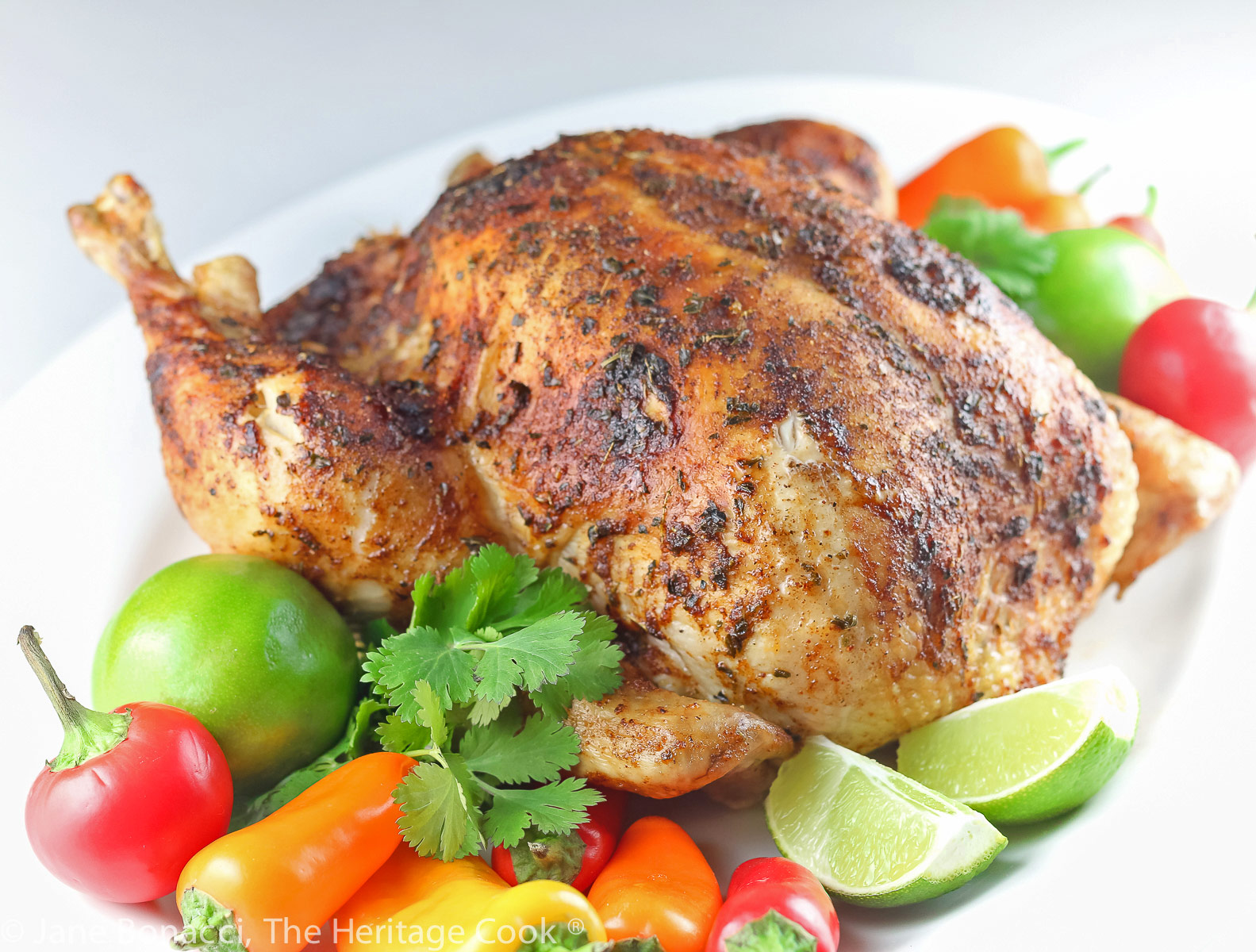 Golden brown Lime Cumin Roast Chicken on a white oval platter, surrounded by mini bell peppers in red, orange, and yellow, cilantro, and limes. It is a joy to make and fills your home with luscious aromas as it cooks © 2023 Jane Bonacci, The Heritage Cook.