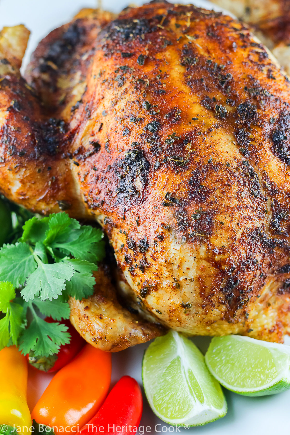 Golden brown Lime Cumin Roast Chicken on a white oval platter, surrounded by mini bell peppers in red, orange, and yellow, cilantro, and limes. It is a joy to make and fills your home with luscious aromas as it cooks © 2023 Jane Bonacci, The Heritage Cook.