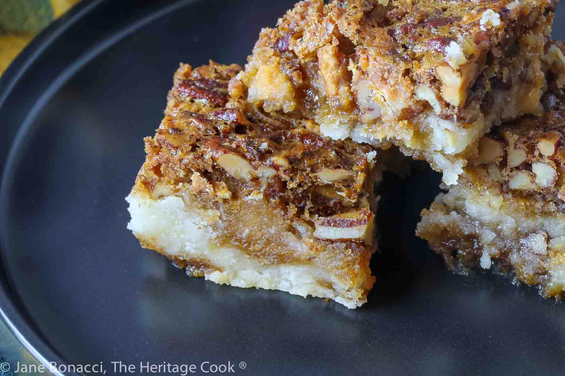 Stack of 4 Pecan Pie Bars on a black plate with a colorful plaid cloth underneath and behind © 2023 Jane Bonacci, The Heritage Cook.