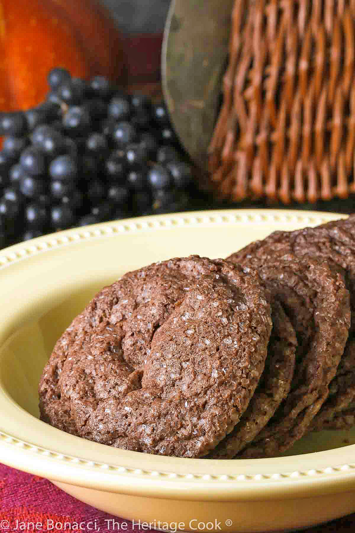 A pile of Sparkling Chocolate Sugar Cookies lined up in a yellow bowl on a colorful tablecloth with pumpkin and grapes behind © 2023 Jane Bonacci, The Heritage Cook. 