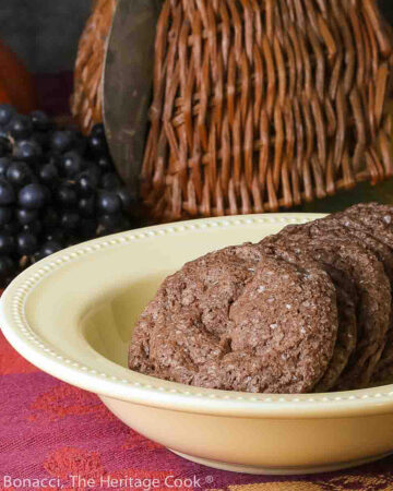 A pile of Sparkling Chocolate Sugar Cookies lined up in a yellow bowl on a colorful tablecloth with pumpkin and grapes behind © 2023 Jane Bonacci, The Heritage Cook.
