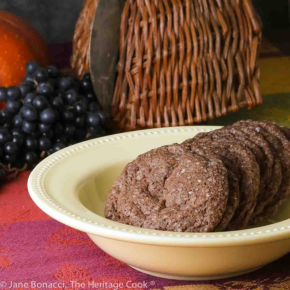 A pile of Sparkling Chocolate Sugar Cookies lined up in a yellow bowl on a colorful tablecloth with pumpkin and grapes behind © 2023 Jane Bonacci, The Heritage Cook.