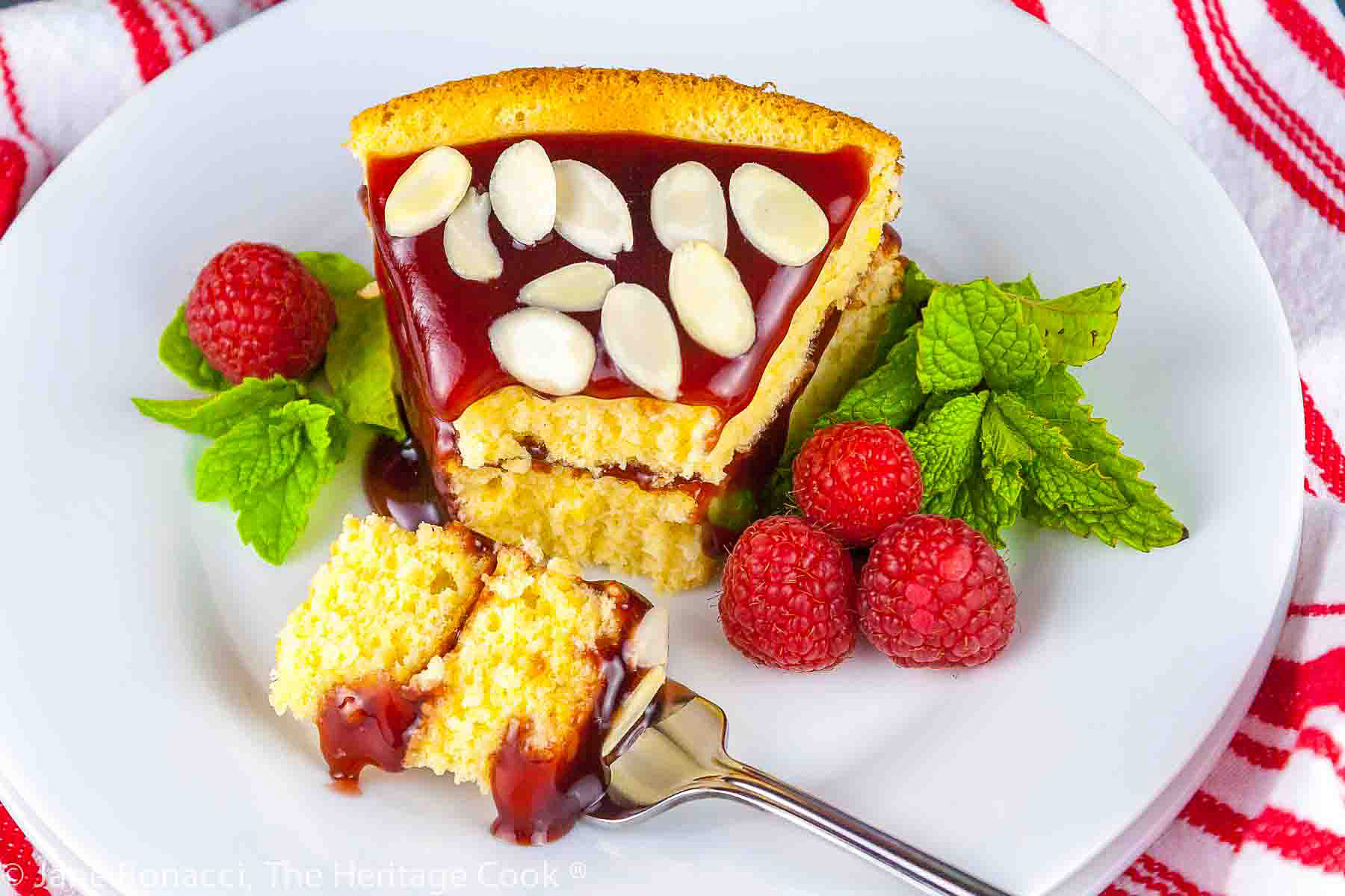 Whole cake and slice of Almond Raspberry Cake with Raspberry-Chocolate-Cassis Glaze on white plate, garnished with mint and fresh raspberries. Some slices topped with sliced almonds © 2023 Jane Bonacci, The Heritage Cook. 