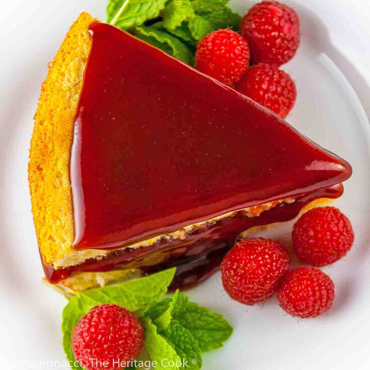 Whole cake and slice of Almond Raspberry Cake with Raspberry-Chocolate-Cassis Glaze on white plate, garnished with mint and fresh raspberries. Some slices topped with sliced almonds © 2023 Jane Bonacci, The Heritage Cook.