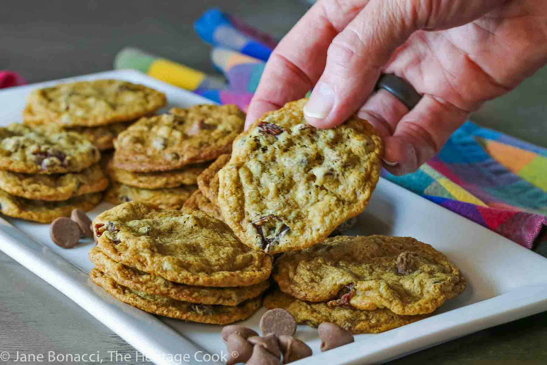 hand stealing a cookie; White rectangular platter filled with stacks of Cherry Chocolate Chip Cookies and scattered chocolate chips with colorful plaid cloths along the sides © 2024 Jane Bonacci, The Heritage Cook. 