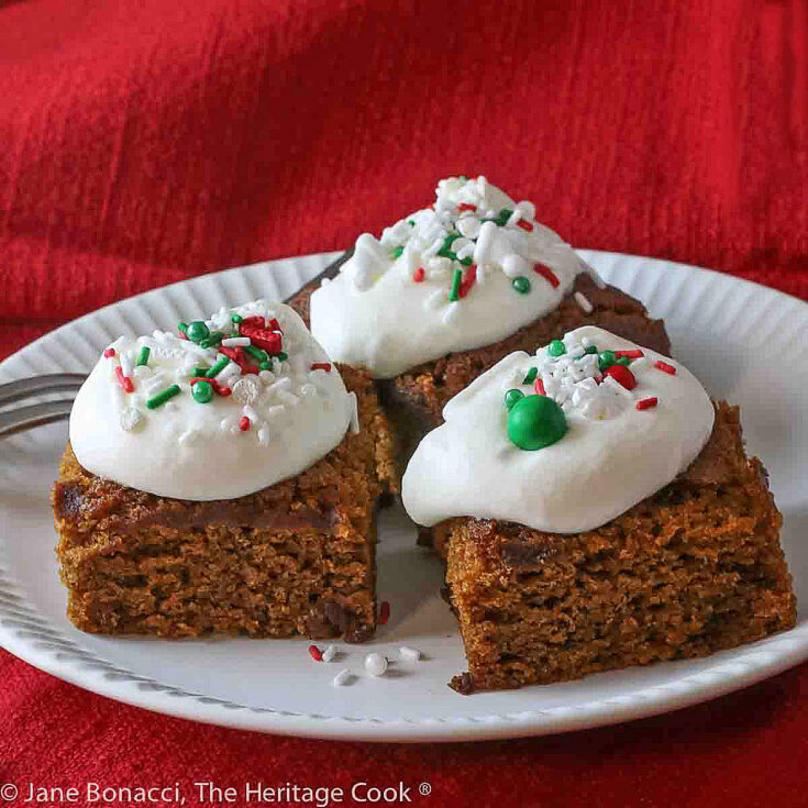 Three pumpkin gingerbread blondies on a white plate with red background linens, topped with a dollop of whipped cream and festive sprinkles © 2023 Jane Bonacci, The Heritage Cook.