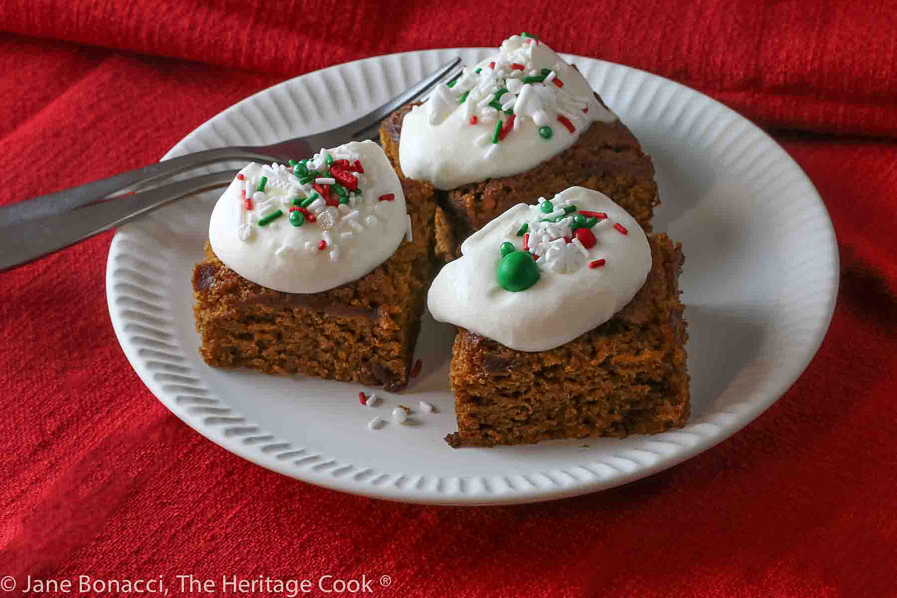 Three pumpkin gingerbread blondies on a white plate with red background linens, topped with a dollop of whipped cream and festive sprinkles © 2023 Jane Bonacci, The Heritage Cook. 