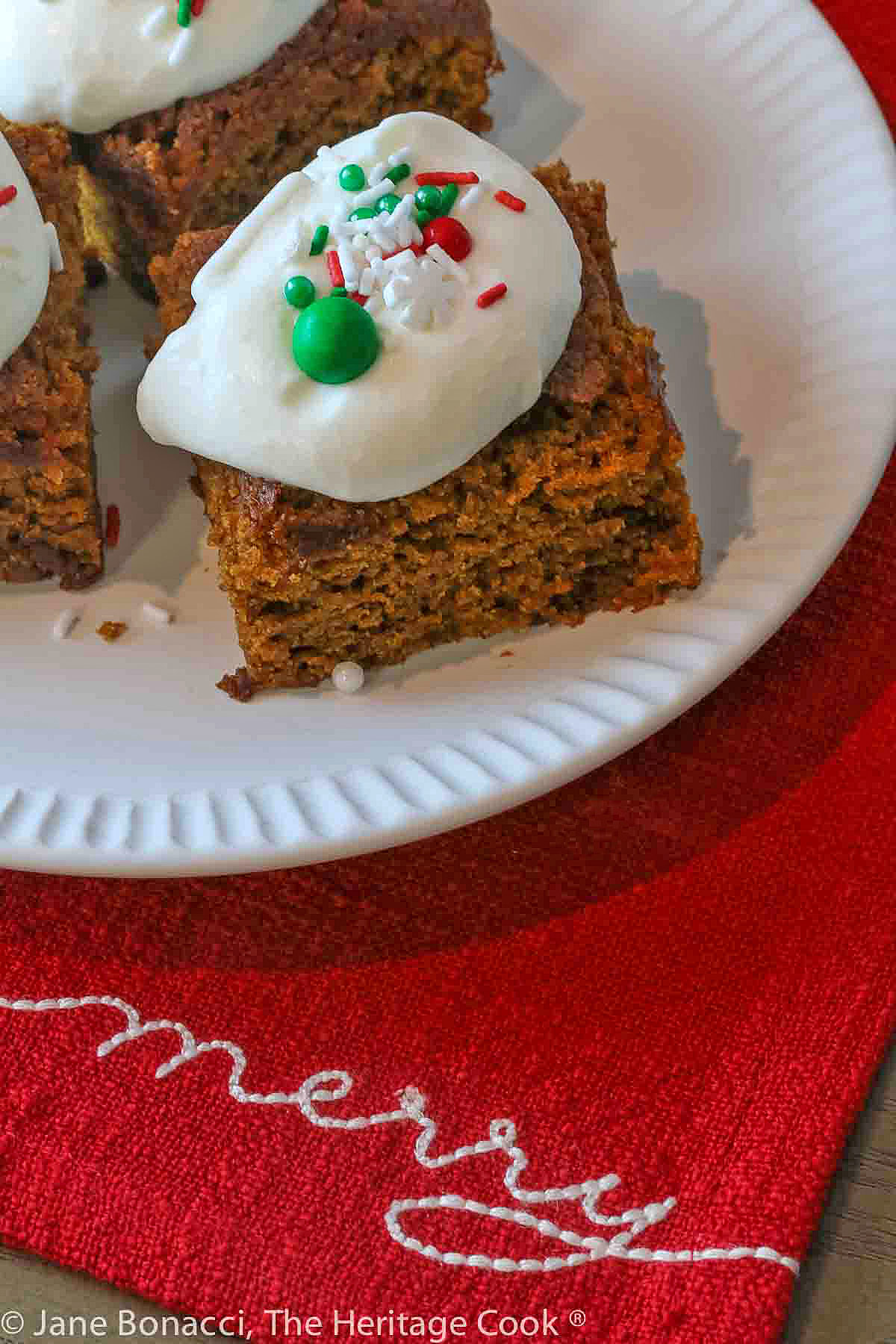 Plate of pumpkin gingerbread blondies on a red cloth embroidered with the word "Merry" on it. 