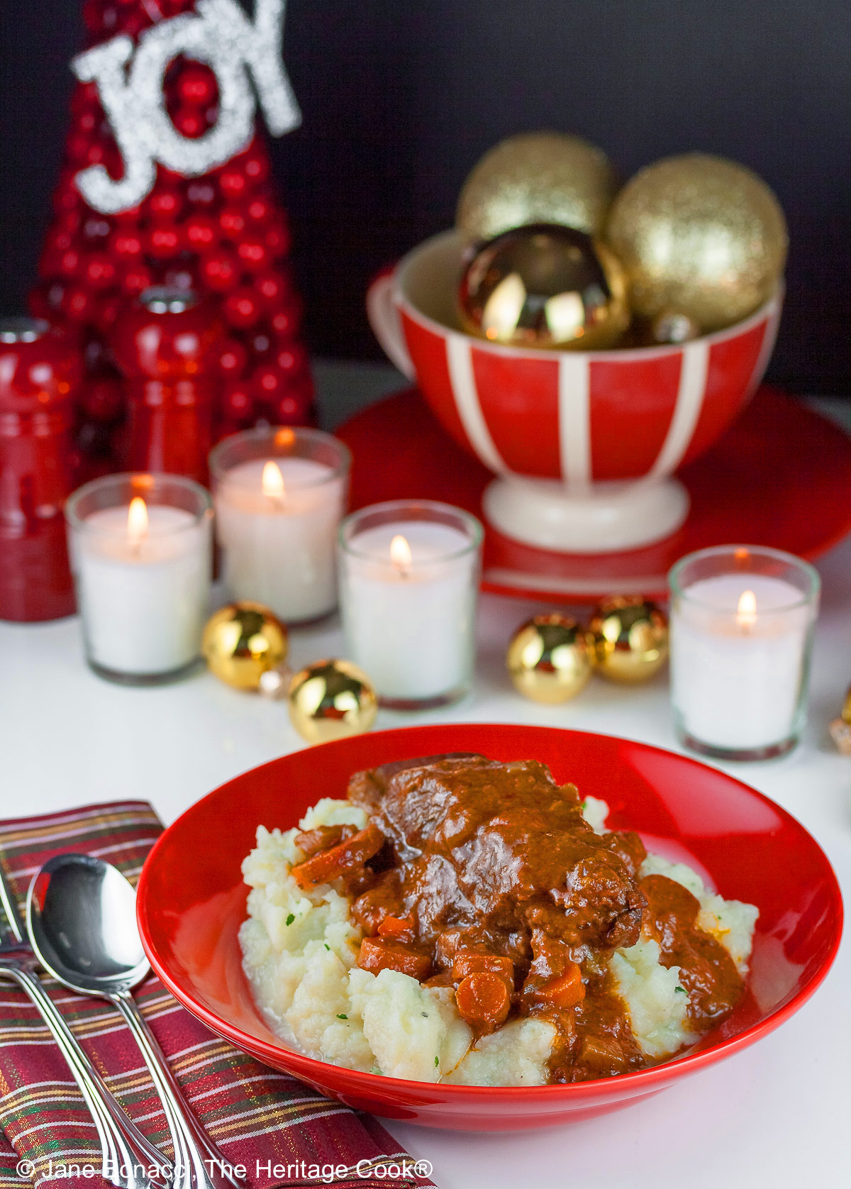 Garlic mashed potatoes lining a red bowl and topped with Red Wine Braised Short Ribs and carrots, in front of festive candles and Christmas decorations with holiday place mats © 2025 Jane Bonacci, The Heritage Cook.