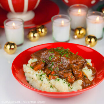 Garlic mashed potatoes lining a red bowl and topped with Red Wine Braised Short Ribs and carrots, in front of festive candles and Christmas decorations with holiday place mats © 2025 Jane Bonacci, The Heritage Cook.