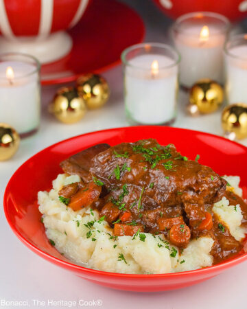 Garlic mashed potatoes lining a red bowl and topped with Red Wine Braised Short Ribs and carrots, in front of festive candles and Christmas decorations with holiday place mats © 2025 Jane Bonacci, The Heritage Cook.