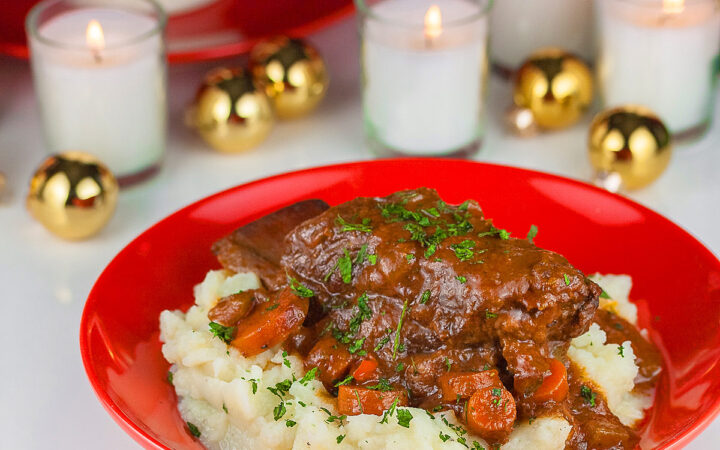 Garlic mashed potatoes lining a red bowl and topped with Red Wine Braised Short Ribs and carrots, in front of festive candles and Christmas decorations with holiday place mats © 2025 Jane Bonacci, The Heritage Cook.