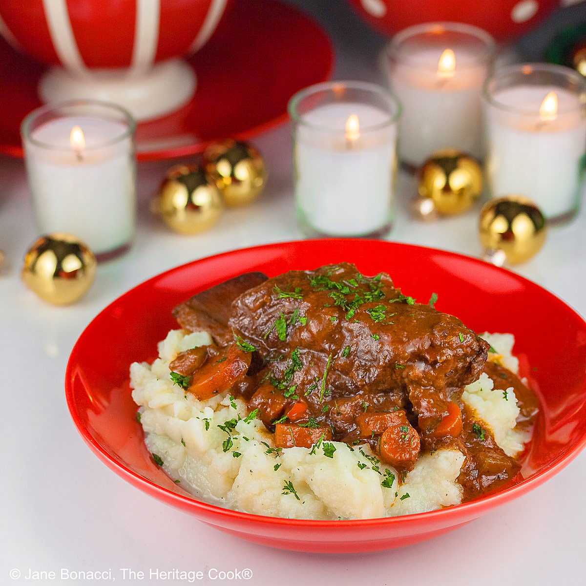 Garlic mashed potatoes lining a red bowl and topped with Red Wine Braised Short Ribs and carrots, in front of festive candles and Christmas decorations with holiday place mats © 2025 Jane Bonacci, The Heritage Cook.