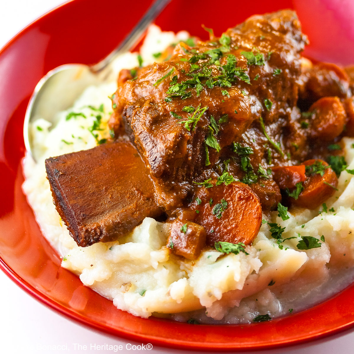 Garlic mashed potatoes lining a red bowl and topped with Red Wine Braised Short Ribs and carrots, in front of festive candles and Christmas decorations with holiday place mats © 2025 Jane Bonacci, The Heritage Cook.