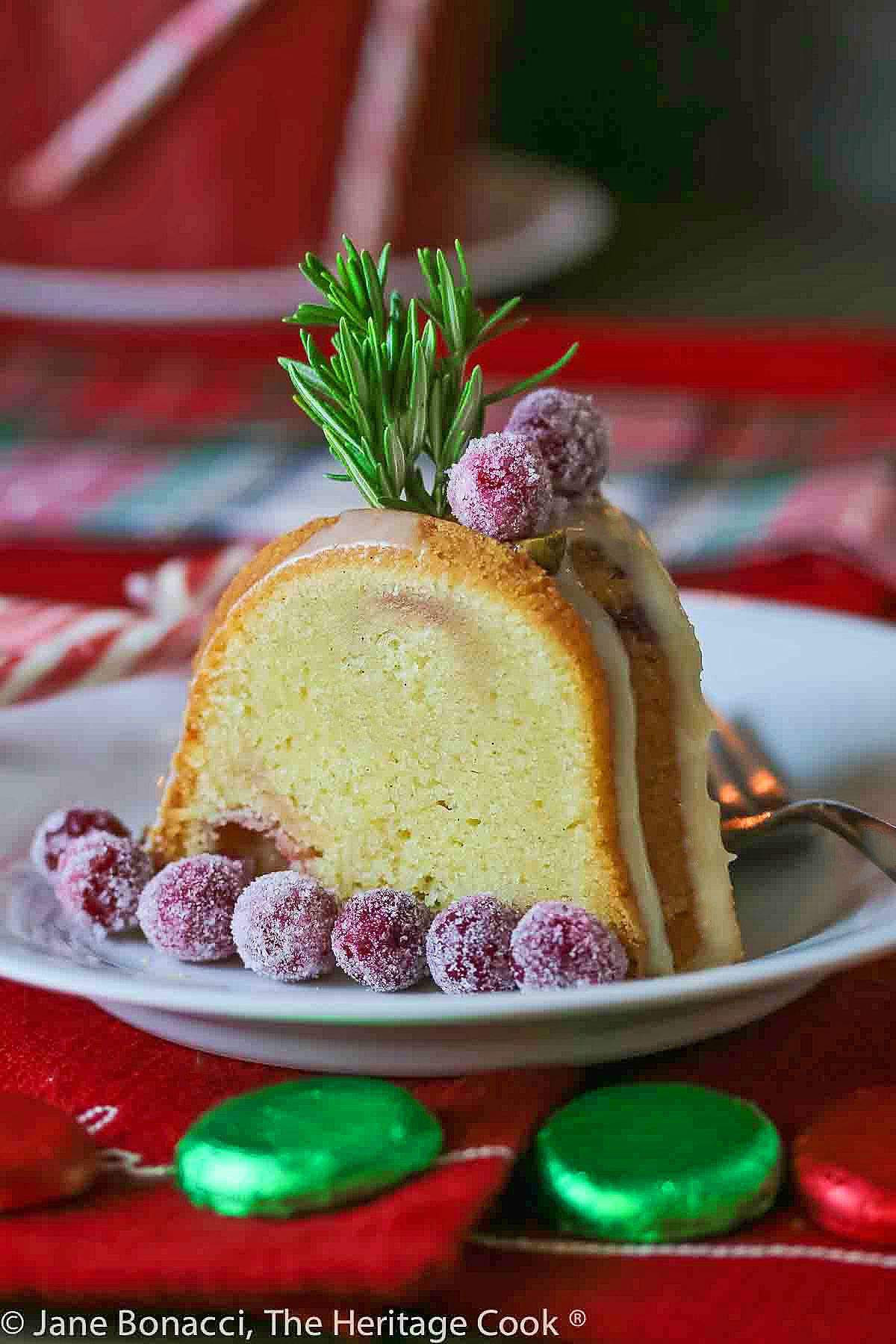 Whole decorated cake with glaze and sugared cranberries and a cut slice on white plates sitting on red cloths with a festive plaid cloth in the back. Some chocolate candies & candy canes around the plates © 2023 Jane Bonacci, The Heritage Cook.