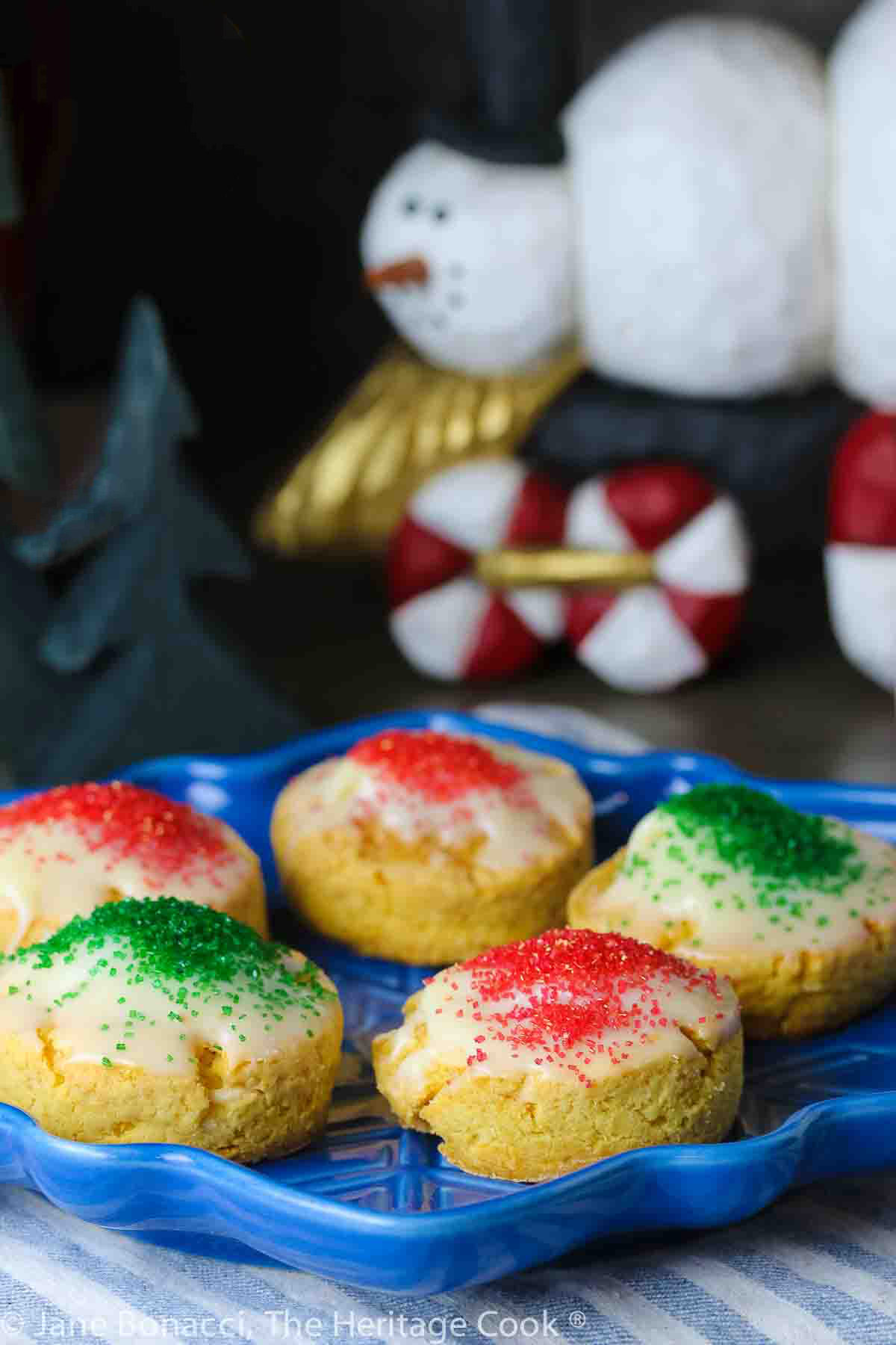 Five White Chocolate Sugared Scones with white chocolate glaze and sprinkled with red and green sugars, served on a blue star-shaped plate on top of a blue and white striped cloth, in front of festive hand-carved snowmen figures © 2023 Jane Bonacci, The Heritage Cook. 