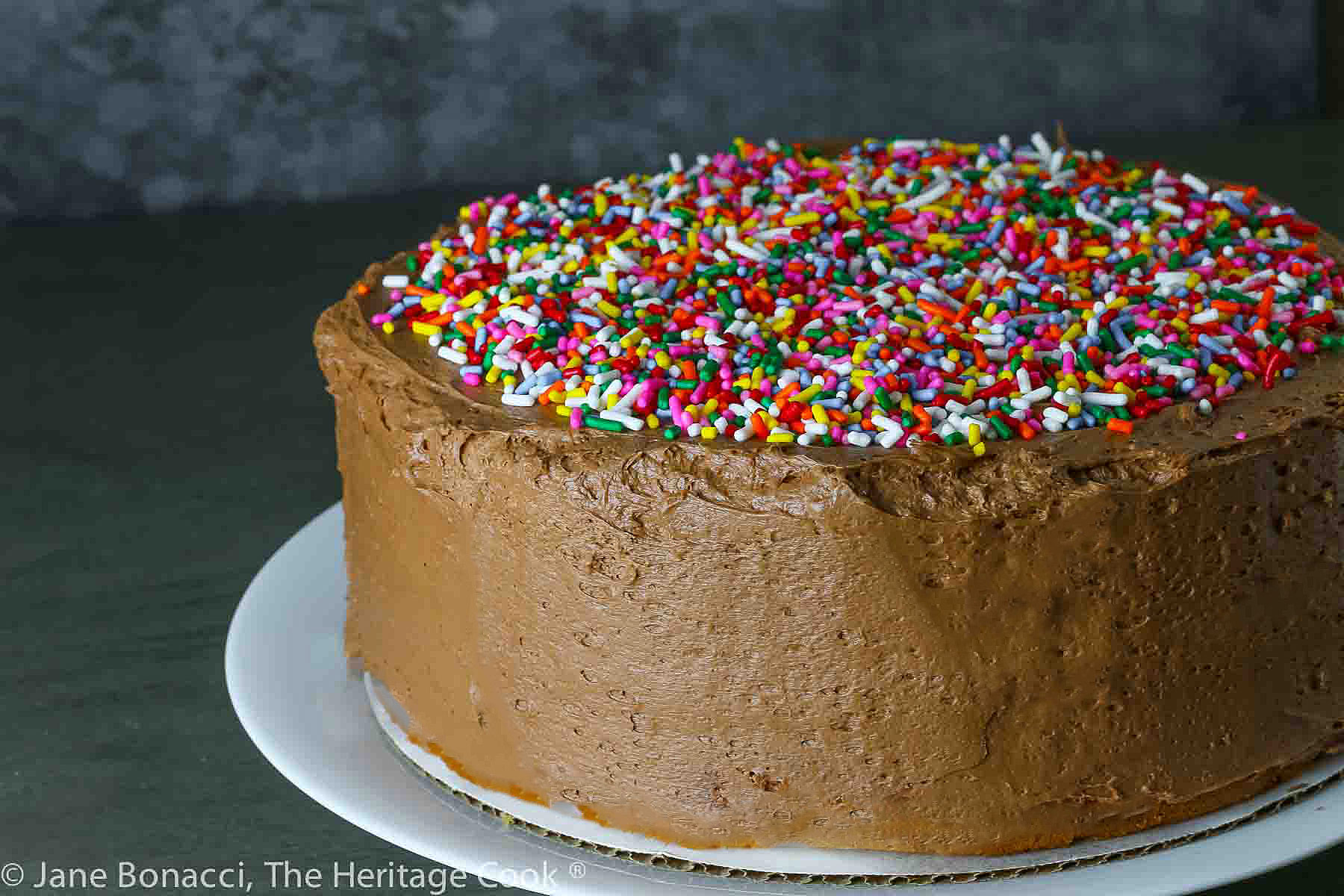 1234 Vanilla Cake with Chocolate Frosting layer cake; slice on a white plate in front of the whole cake with slices cut out of it; covered in colorful sprinkles on the top and a glass of milk behind it; © 2024 Jane Bonacci, The Heritage Cook.