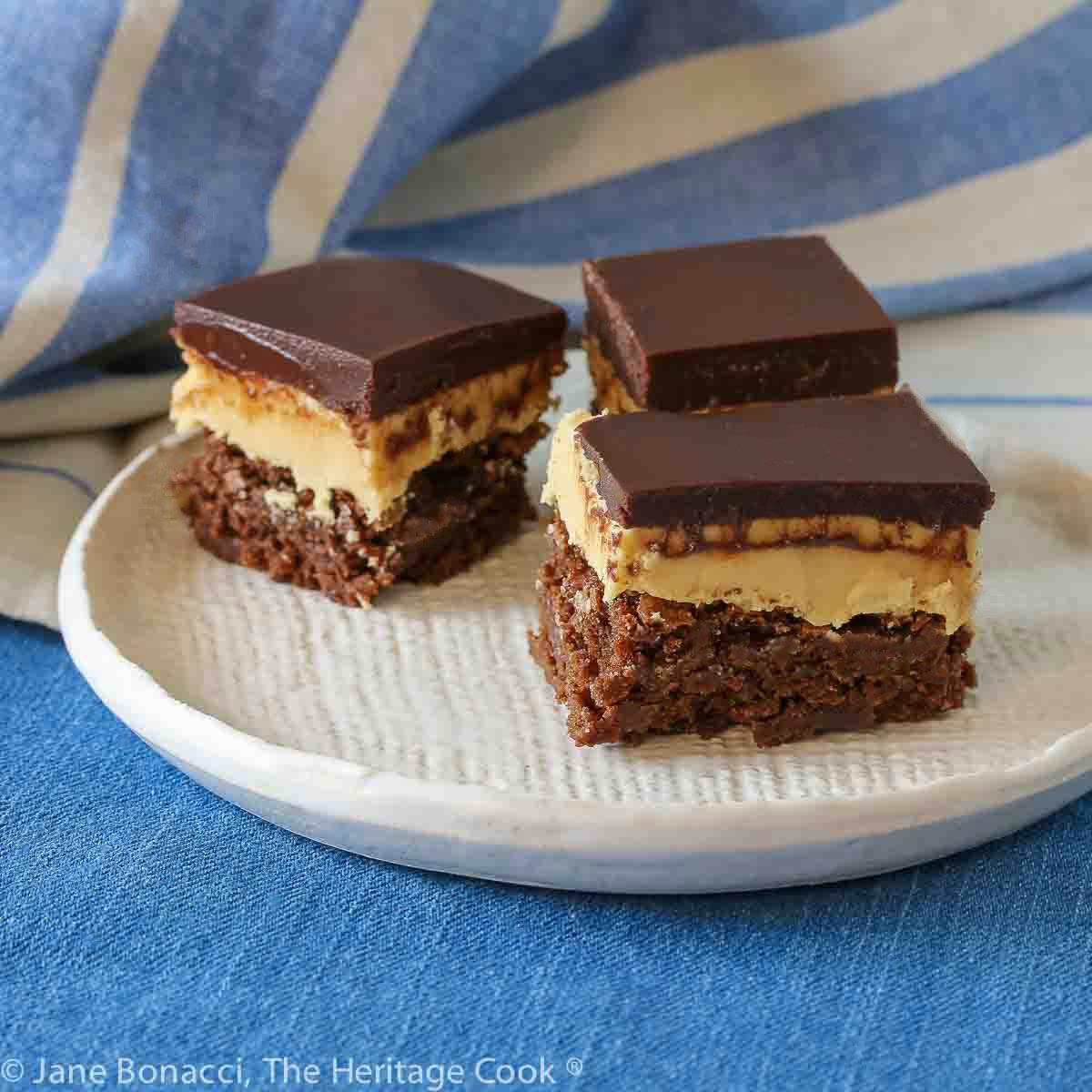 Three squares of layered Buckeye Peanut Butter Brownies on a white etched pottery plate set on a blue denim cloth with a blue striped cloth behind © 2024 Jane Bonacci, The Heritage Cook. 