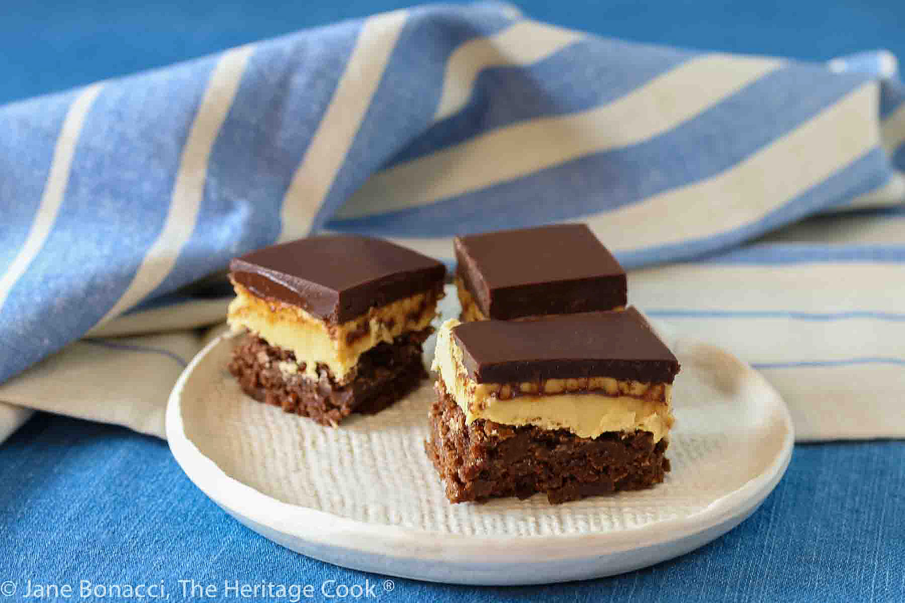 Three squares of layered Buckeye Peanut Butter Brownies on a white etched pottery plate set on a blue denim cloth with a blue striped cloth behind © 2024 Jane Bonacci, The Heritage Cook. 
