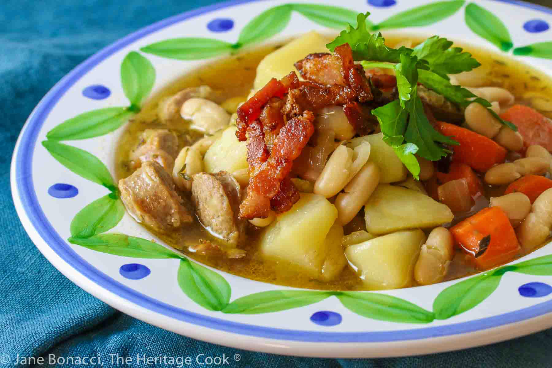 French Cassoulet Soup with sausages, beans, potatoes, duck, and rich broth served in a shallow bowl with a big painted rim, piled high with crispy bacon and a parsley sprig © 2024 Jane Bonacci, The Heritage Cook. 