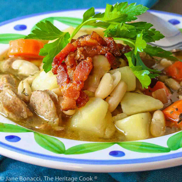French Cassoulet Soup with sausages, beans, potatoes, duck, and rich broth served in a shallow bowl with a big painted rim, piled high with crispy bacon and a parsley sprig © 2024 Jane Bonacci, The Heritage Cook.