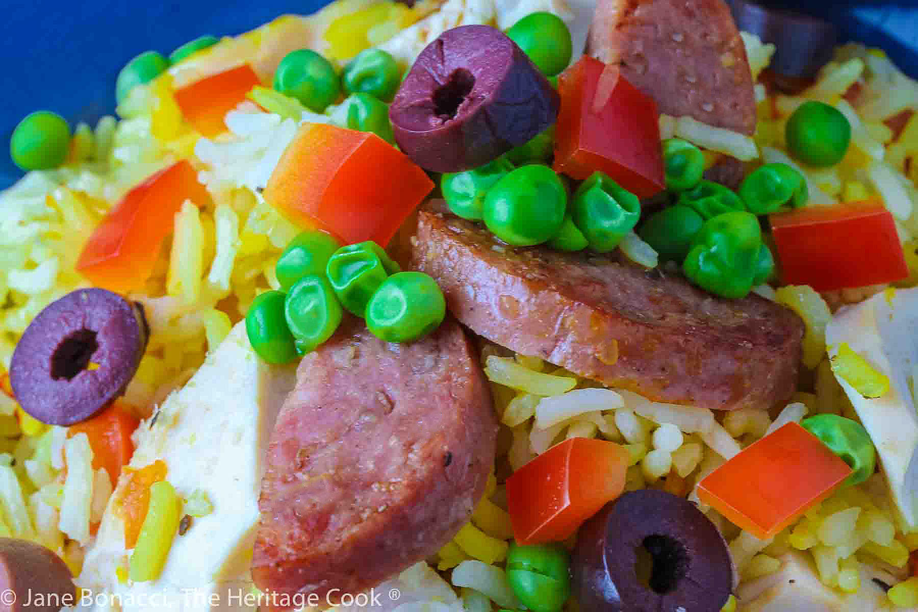Portuguese Chicken and Rice in a dark blue bowl, with sliced chicken, sausage, and saffron rice, topped with kalamata olives, red bell pepper, and green peas © 2024 Jane Bonacci, The Heritage Cook. 