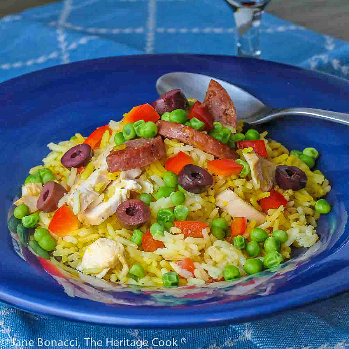 Portuguese Chicken and Rice in a dark blue bowl, with sliced chicken, sausage, and saffron rice, topped with kalamata olives, red bell pepper, and green peas © 2024 Jane Bonacci, The Heritage Cook.