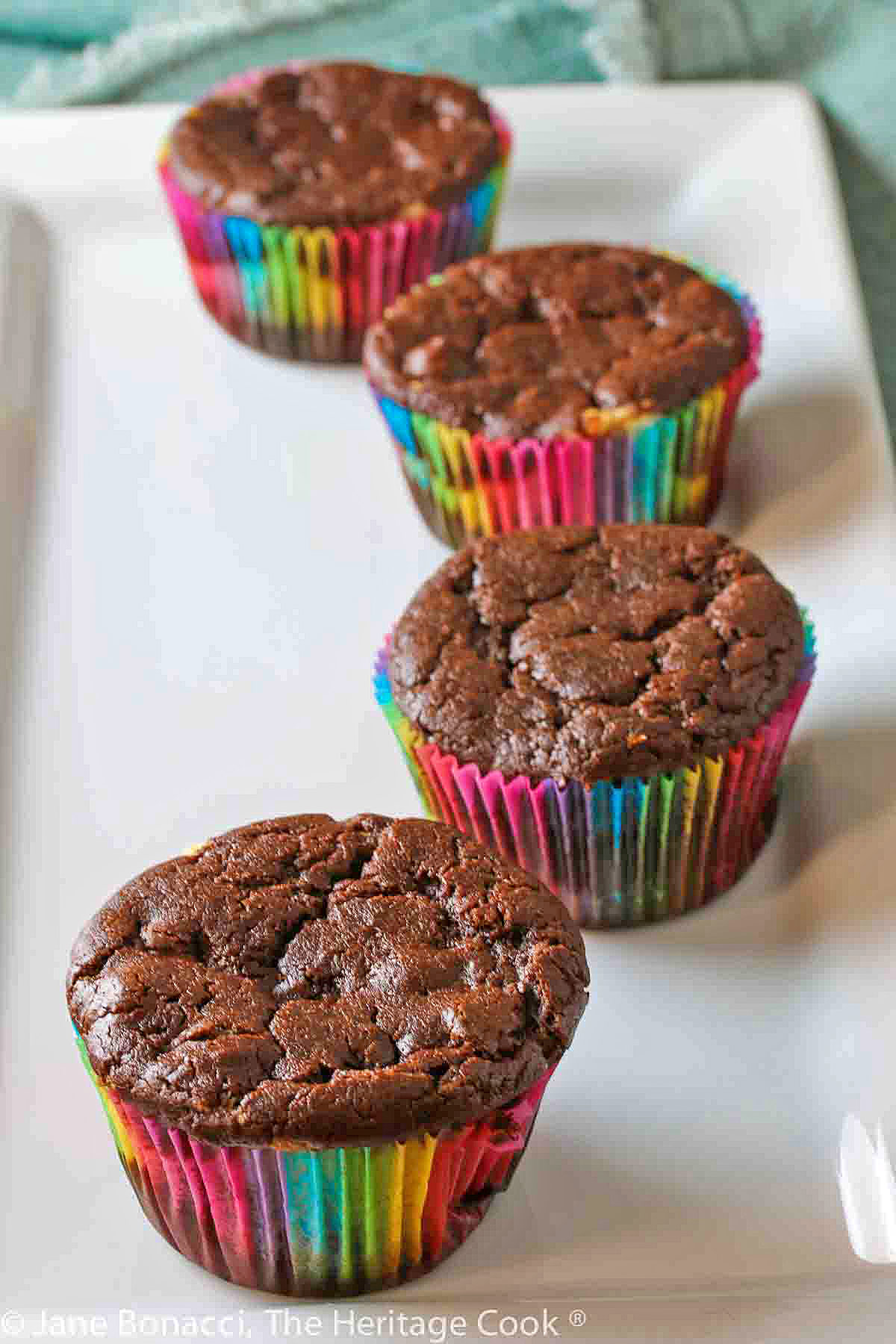 Chocolate muffins baked in rainbow colored paper liners, lined up on a white rectangular platter © 2024 Jane Bonacci, The Heritage Cook. 