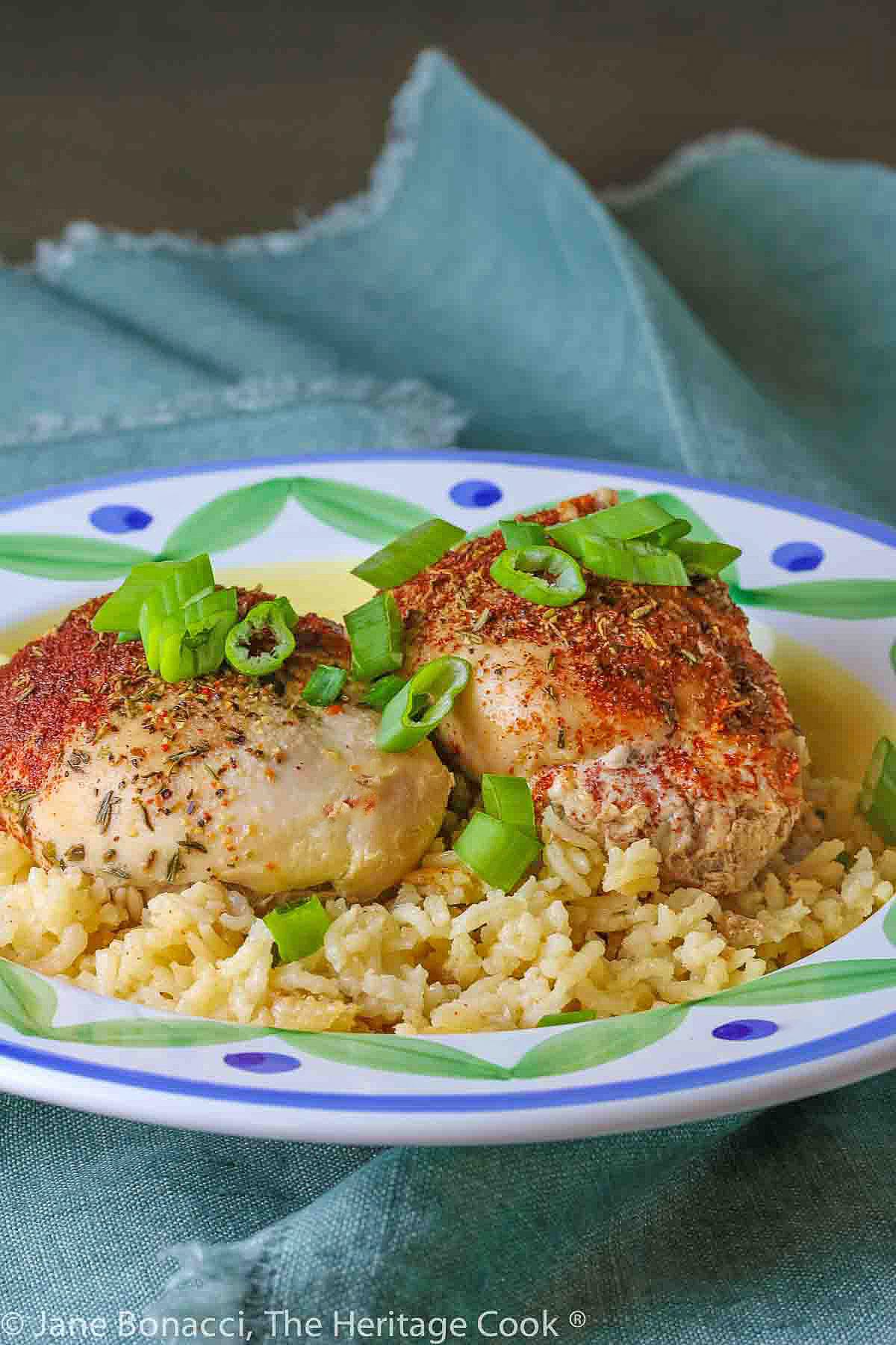 Two rounded chicken thighs sprinkled with paprika and seasonings resting in a bowl of rice with sliced green onions over the top © 2024 Jane Bonacci, The Heritage Cook. 