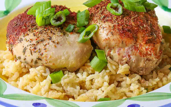 Two rounded chicken thighs sprinkled with paprika and seasonings resting in a bowl of rice with sliced green onions over the top © 2024 Jane Bonacci, The Heritage Cook.