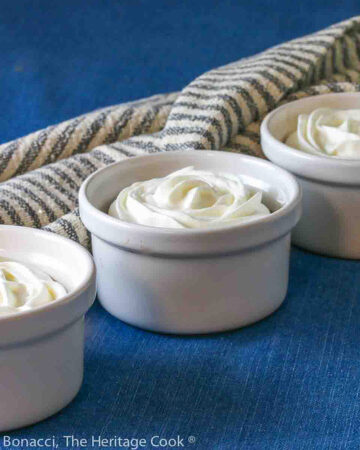 White ramekins filled with dark brown Easy Creamy Chocolate Pudding, topped with beautifully piped white whipped cream roses on a blue background with a grey and white stripped cloth © 2024 Jane Bonacci, The Heritage Cook.