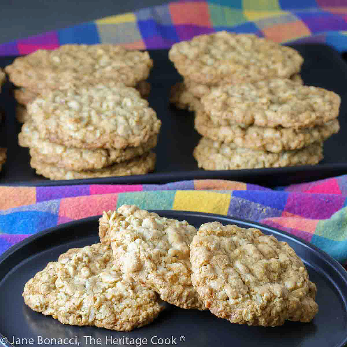 White Chocolate Pecan Oatmeal Cookies stacked on a black rectangular platter some with a small black plate in front with 3 cookies on it and brightly colored plaid cloths around © 2024 Jane Bonacci, The Heritage Cook.