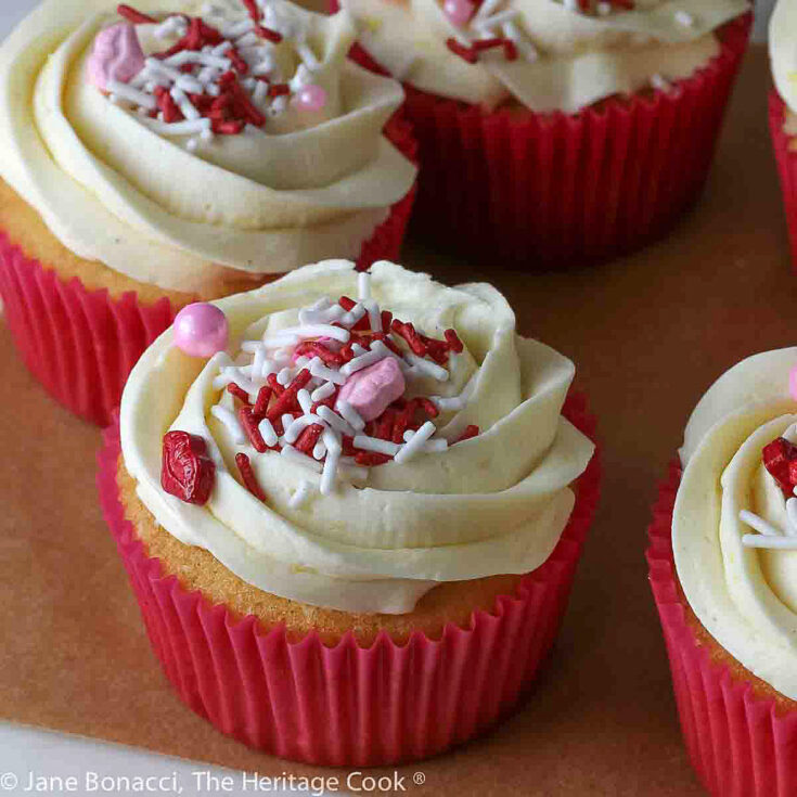 Multiple vanilla cupcakes in red paper liners, filled with whipped white chocolate ganache, topped with lemon kissed buttercream frosting and sprinkled with festive sprinkles © 2024 Jane Bonacci, The Heritage Cook.